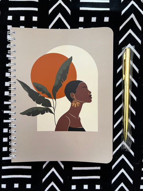 Product photo of spiral bound notebook with image of black woman gazing confidently upward in sky with autumn sun. Shown with gold pen on African mud cloth tribal background