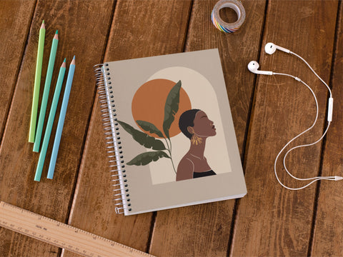 Product photo of a spiral bound notebook with image of black woman gazing confidently upward in sky with autumn sun. Shown on wooden desk background